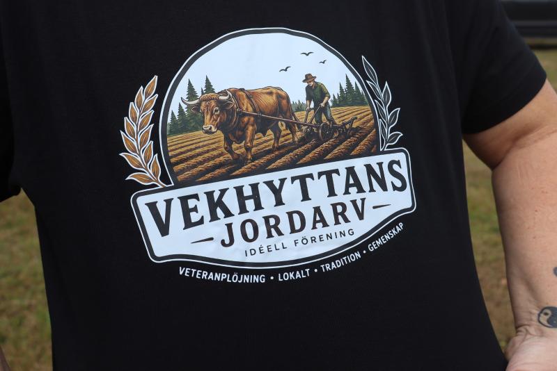 En person bär en svart tröja med en logotyp som föreställer en bonde som plöjer en åker med oxar och texten ”Vekhyttans Jordarv Idéell Förening”.