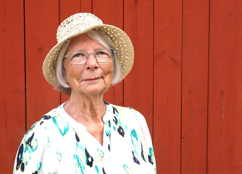 En äldre kvinna med glasögon och en stråhatt står framför en röd trävägg, klädd i en vit mönstrad blus.