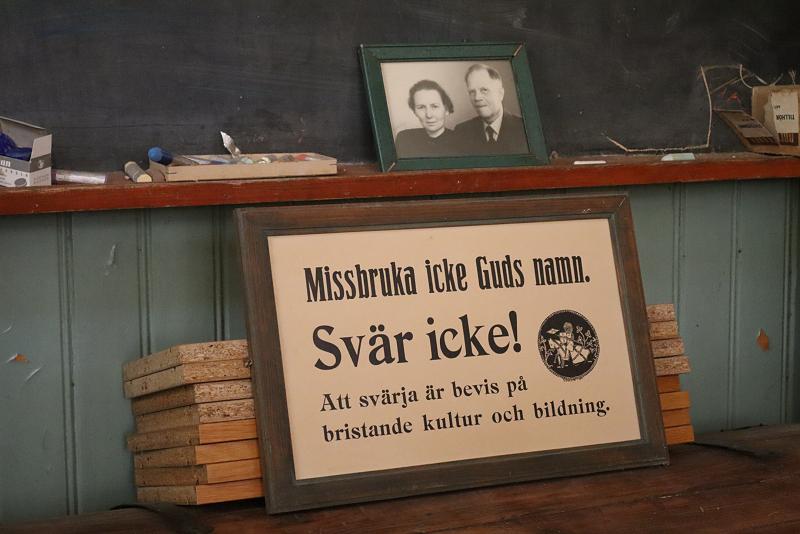 Ett inramat foto och en skylt på svenska på en trähylla, med krita och böcker i närheten; skylten varnar för att missbruka Guds namn och svära.