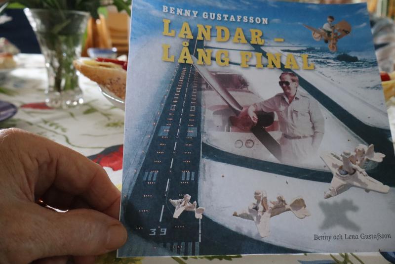 En hand håller en bok med titeln ”Landar – Lång Final” av Benny Gustafsson, där omslaget visar en flygplatslandningsbana, en man med solglasögon och fallskärmshoppare.