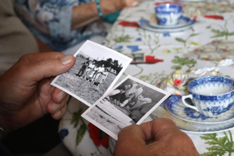 En person håller upp två svartvita fotografier ovanför en blommig duk med tekoppar och fat, medan en annan person sitter bredvid.