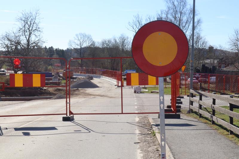 En väg är avstängd med röda metallbarriärer och en rund trafikskylt med förbud mot infart; byggmaterial och utrustning syns bakom barriären.