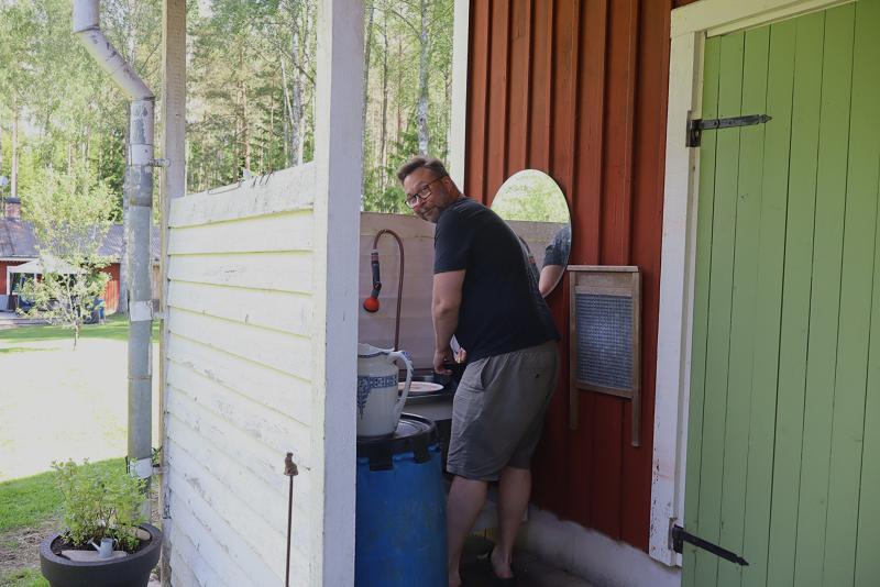 En person står på en veranda utanför en röd träbyggnad och diskar vid ett handfat bredvid en spegel och en tvättbräda. Träd och en gräsmatta syns i bakgrunden.