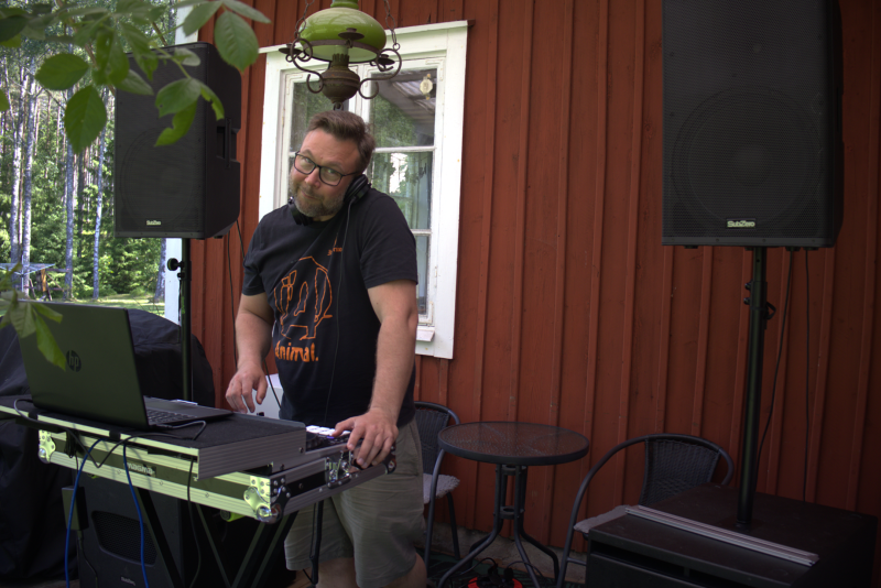 En man står vid ett DJ-bås med en bärbar dator och ljudutrustning utomhus, bredvid en röd träbyggnad med stora högtalare och ett fönster i bakgrunden.