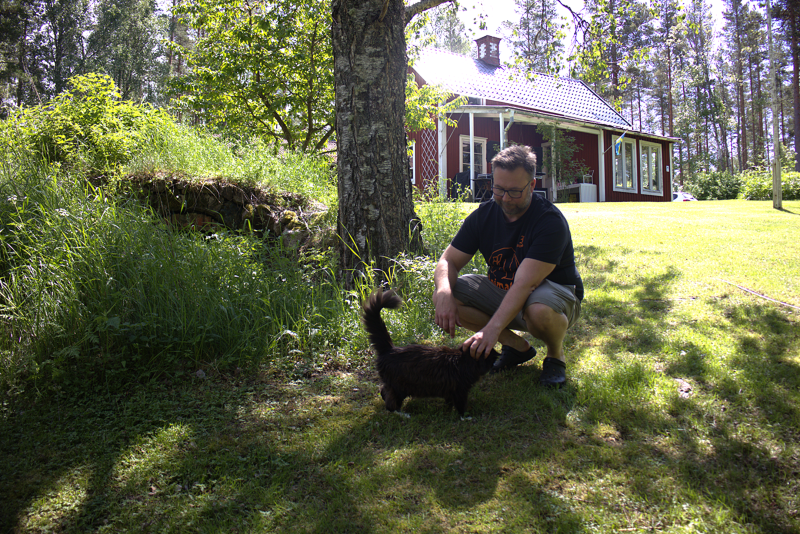 En man klappar en svart katt under ett träd i en solig trädgård, med ett rött hus och gröna bladverk i bakgrunden.