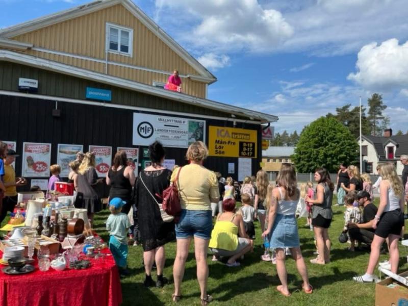 En folkmassa samlas utanför en gul byggnad med skyltar, vänd mot en person på taket. På bord i gräsytan finns olika föremål till försäljning.