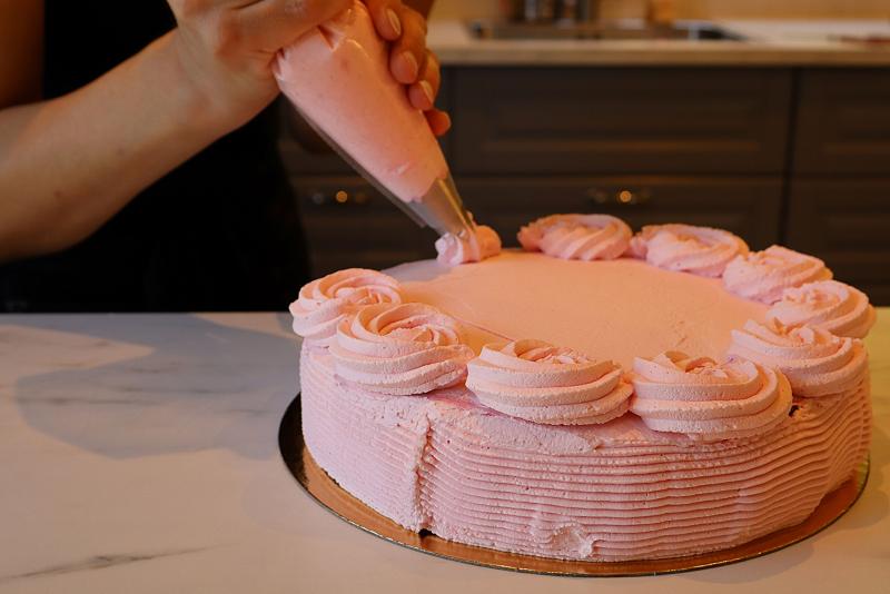 En person dekorerar en rund tårta med rosa glasyr och spritsar rosetter runt kanten på en bänkskiva i marmor.