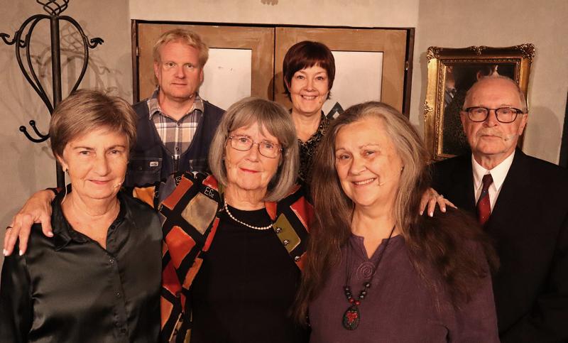 Sex äldre vuxna, tre män och tre kvinnor, står tillsammans inomhus och poserar för ett gruppfoto framför en vägg med inramad konst och en klädhängare.