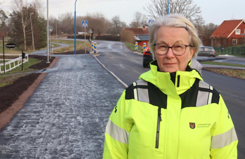 En äldre kvinna iförd varseljacka står vid sidan av en delvis renoverad väg i ett förortsområde med hus och träd i bakgrunden.