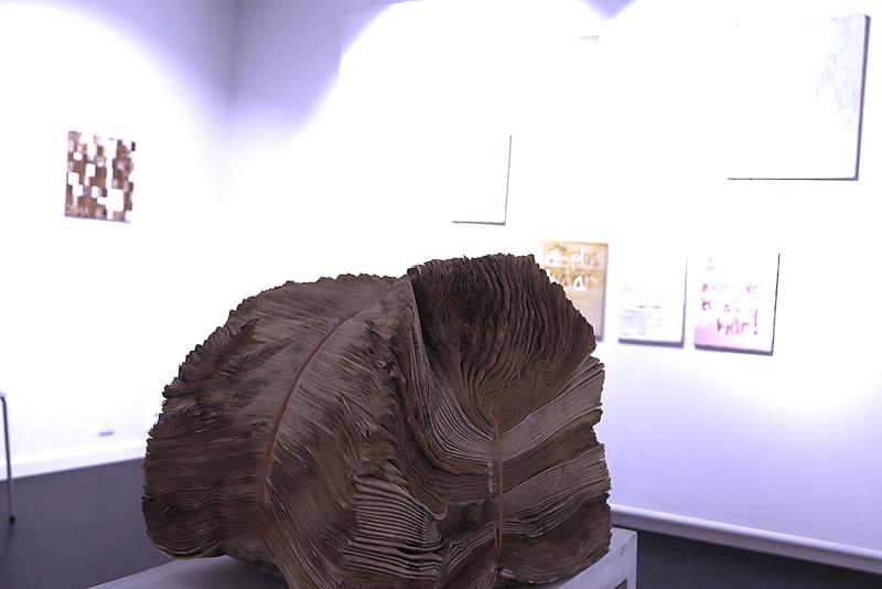 En stor, sliten bok med skrynkliga sidor visas på en piedestal i ett konstgalleri, med inramade konstverk hängande på de vita väggarna i bakgrunden.