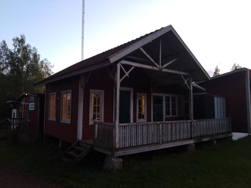 Ett trähus med sadeltak och en omslutande veranda. Fönstren är upplysta inifrån, och det finns träd i bakgrunden.