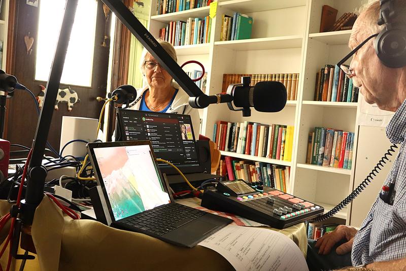 Två personer med hörlurar på sig sitter vid ett bord med mikrofoner, en bärbar dator, ljudutrustning och papper i ett rum med bokhyllor.