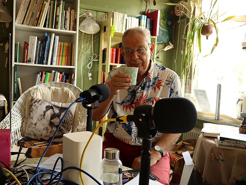 En äldre man i blommig skjorta sitter vid ett bord med mikrofoner och håller i en kopp, omgiven av böcker och växter i ett mysigt rum.