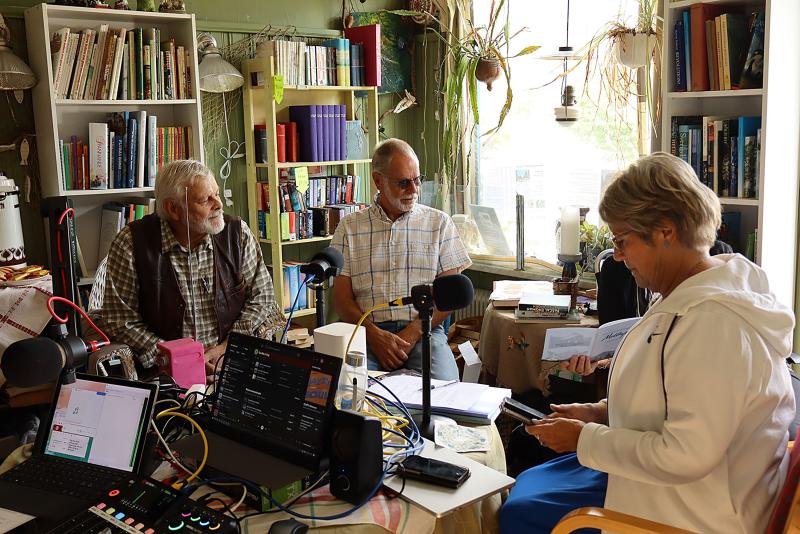 Tre äldre personer sitter runt ett bord med mikrofoner och ljudutrustning och för en diskussion i ett rum med bokhyllor, växter och naturligt ljus från ett fönster.