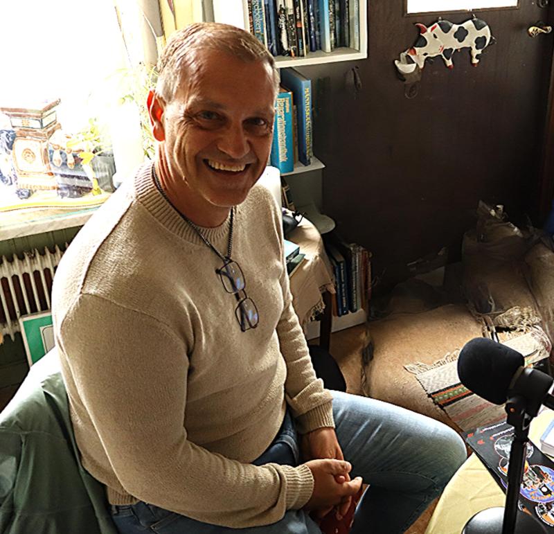 En man i ljus tröja och glasögon sitter i ett mysigt, rörigt rum nära en mikrofon och ler mot kameran.