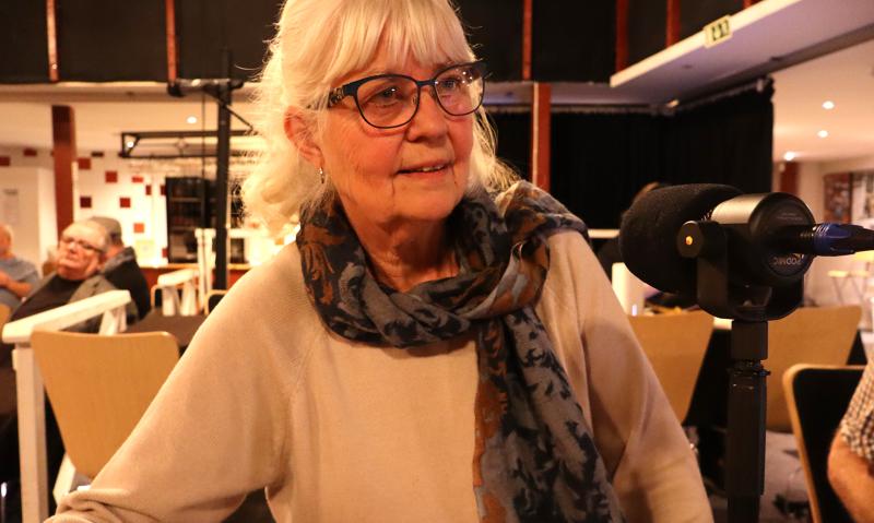 En äldre kvinna med glasögon och mönstrad scarf sitter vid en mikrofon i ett väl upplyst rum med andra människor och stolar i bakgrunden.