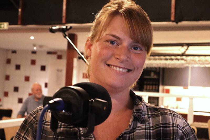 En kvinna med blont hår och rutig skjorta ler mot kameran när hon står framför en mikrofon i en inomhusmiljö. En man syns i den suddiga bakgrunden.