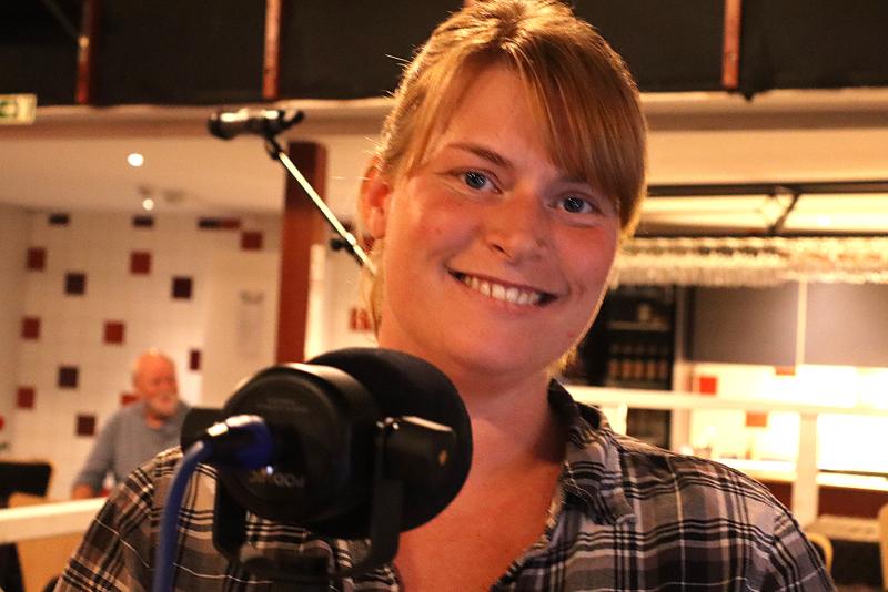 En kvinna med blont hår och rutig skjorta står framför en mikrofon och ler. Bakgrunden ser ut att vara en inomhusmiljö med en sittande person synlig.