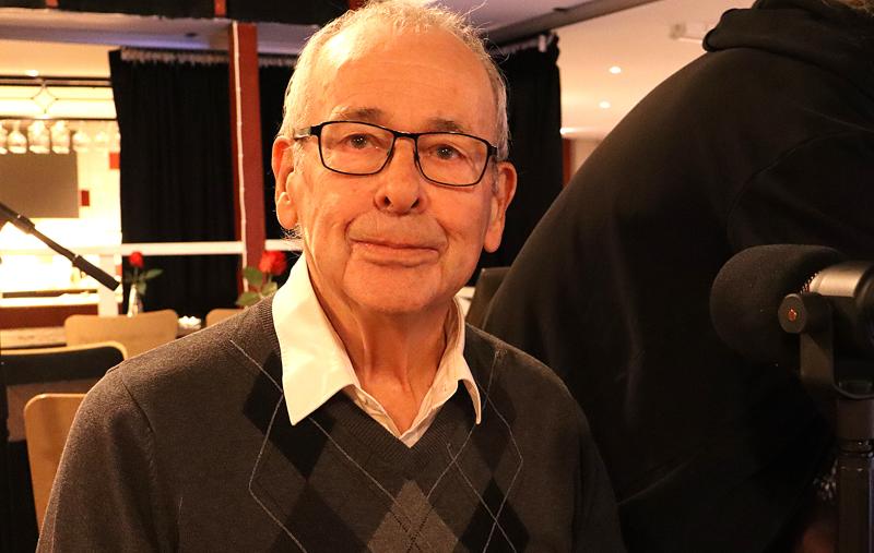 En äldre man med glasögon och en grå argyle-tröja sitter inomhus vid ett bord och tittar in i kameran. Bredvid honom finns en mikrofon och en annan person som är delvis synlig.