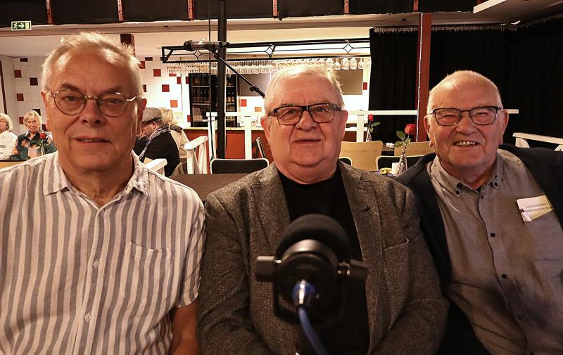 Tre äldre män sitter sida vid sida vid ett bord i ett väl upplyst rum, vända mot kameran. En mikrofon syns i förgrunden. Andra människor och bord finns i bakgrunden.