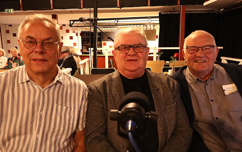 Tre äldre män sitter sida vid sida inomhus, vända mot kameran, med en mikrofon framför sig och människor sittande i bakgrunden.
