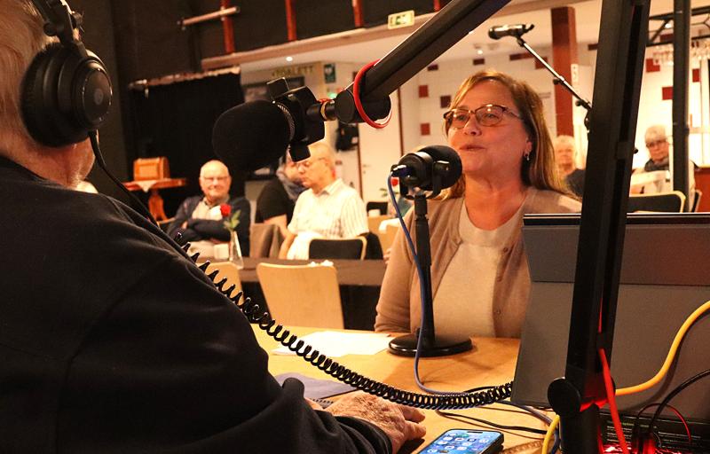 En kvinna talar i en mikrofon under en direktsänd radiointervju inför publik. Ljudutrustning och kablar syns på bordet.