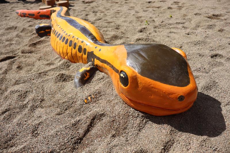 En stor, orange och svart salamanderskulptur ligger på sandig mark och har framträdande ögon och ett brett huvud.