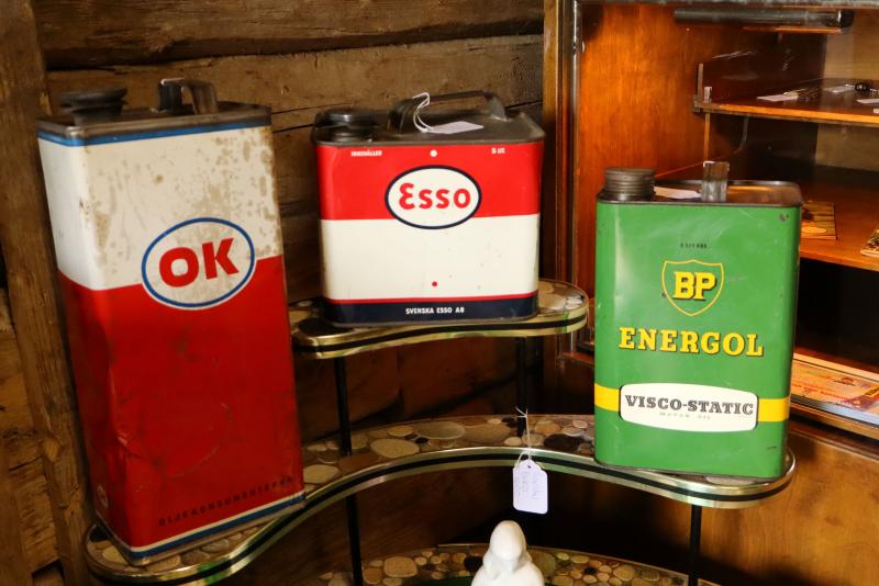 Tre gamla oljecisterner med märkena OK, Esso och BP Energol står på glashyllor framför en trävägg.