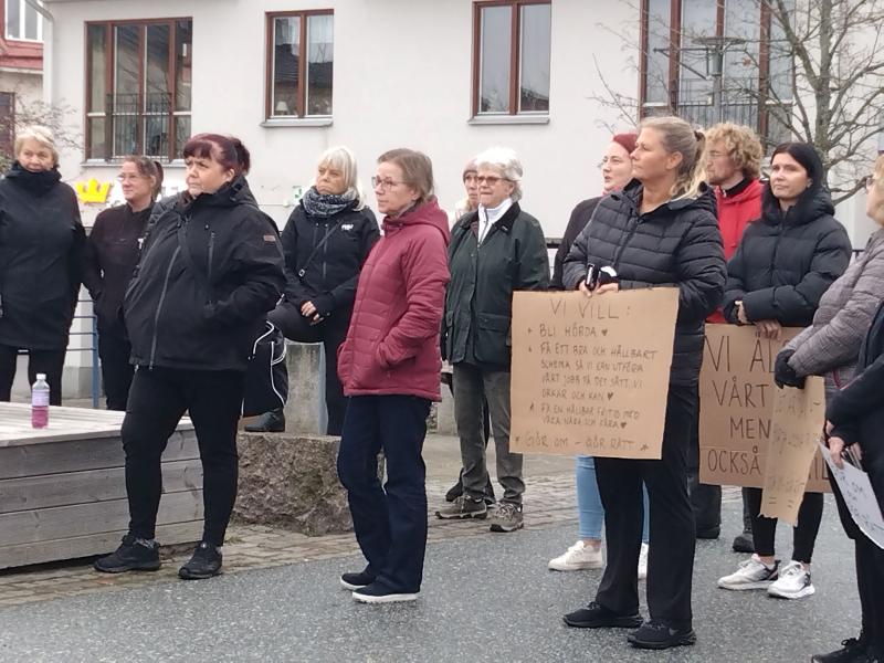 En grupp människor står utomhus och håller i skyltar med text. Vissa är klädda i jackor, vilket tyder på kallare väder. En byggnad syns i bakgrunden.