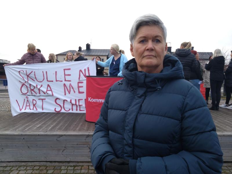 En person i blå jacka står i förgrunden. I bakgrunden håller människor en skylt där det står 