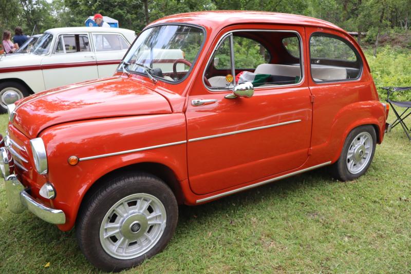 En röd veteranbil av äldre modell står parkerad på gräset vid ett utomhusevenemang, med en annan veteranbil och träd synliga i bakgrunden.