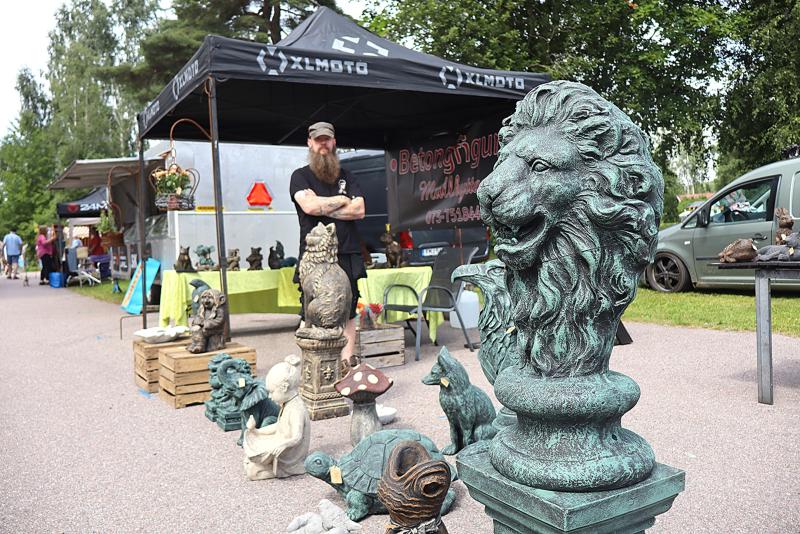 En marknadsbod utomhus där olika trädgårdsstatyer ställs ut, bland annat en stor skulptur föreställande ett lejonhuvud i förgrunden, med en person som står bakom bordet.