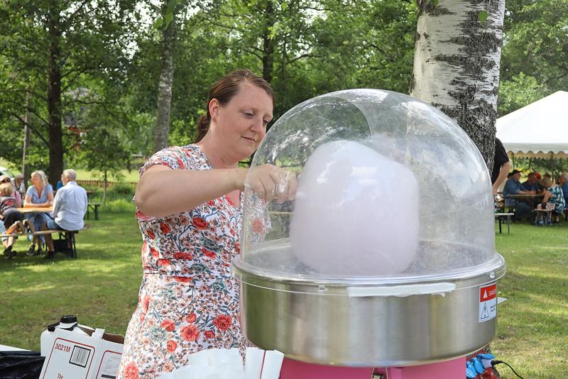 En kvinna i en blommig klänning gör sockervadd utomhus vid ett evenemang i parken, med människor som sitter vid picknickbord i bakgrunden.
