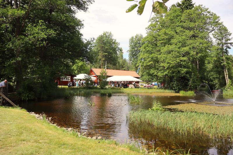 En liten damm med en fontän, omgiven av gräs och träd, med en röd byggnad och människor som samlats under tält i bakgrunden.