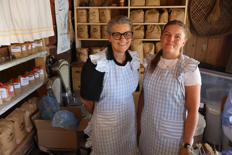 Två kvinnor i blå rutiga förkläden står i en butik fylld med hyllor av förpackade varor. Båda ler, och miljön ger ett välorganiserat och rustikt intryck.