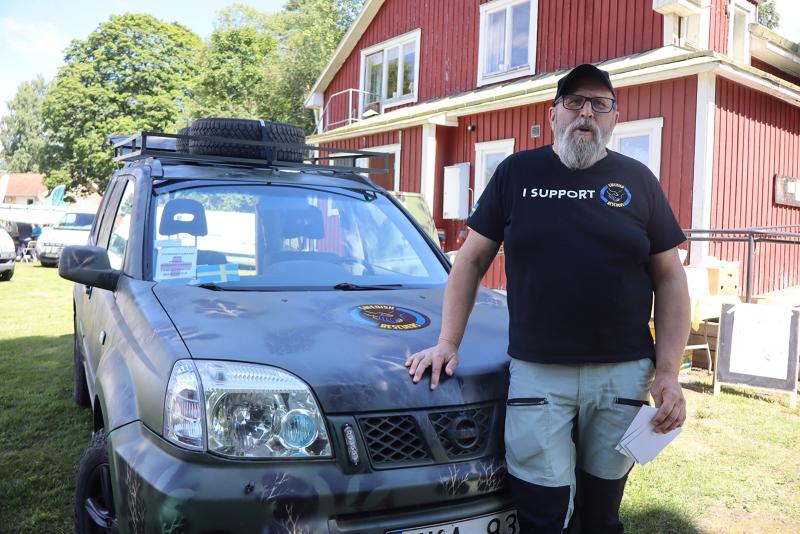 En man med glasögon och en T-shirt med texten ”I SUPPORT” står bredvid en kamouflerad SUV och håller i några papper framför ett rött trähus en solig dag.