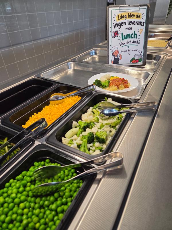 En serveringslinje i en cafeteria med brickor med ärtor, majs, blandade grönsaker och en tallrik med mat. En skylt på svenska förklarar att leveransen uteblev, men att lunchen ändå serveras.