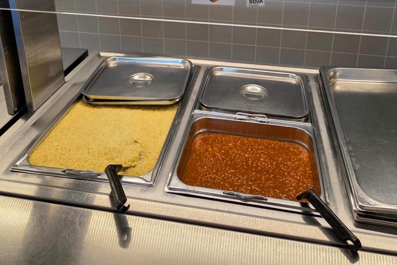 Två metallbrickor på en cafeteriadisk, den ena fylld med tillagad couscous och den andra med en röd sås med köttfärs, båda delvis täckta med lock.