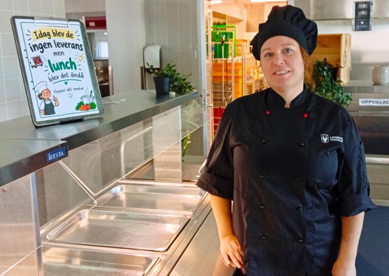 En kvinna i kockuniform står bredvid en tom serveringsdisk med en skylt på svenska där det står att ingen leverans har skett, men att lunchen ändå har tillagats.