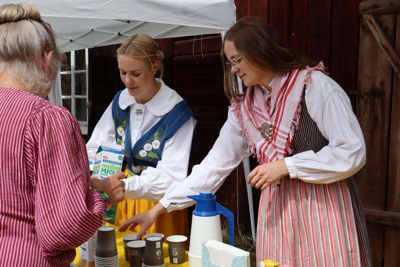 Två kvinnor i traditionella kläder serverar drycker till en kvinna vid ett bord utomhus med muggar, kartonger, termos och servetter.
