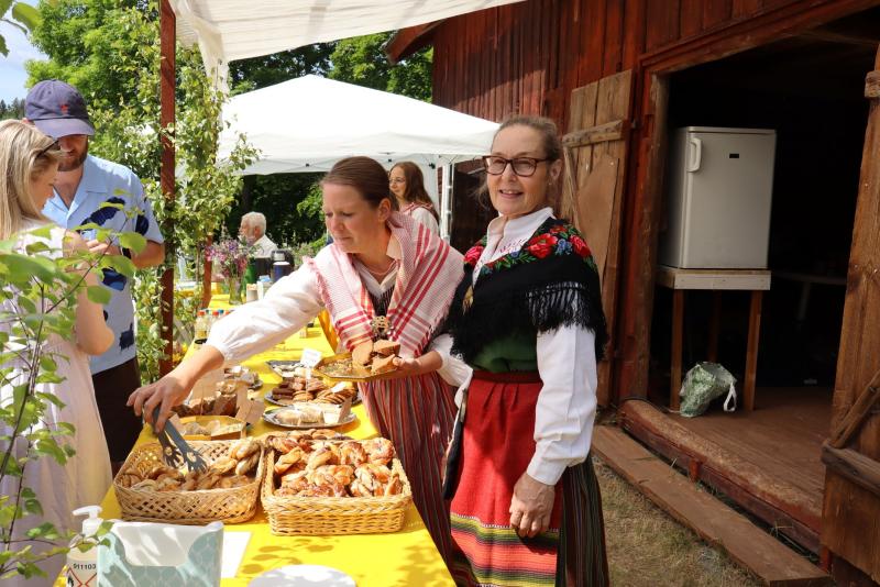 Flera personer i traditionella kläder står vid ett bord med olika bakverk och mat utomhus bredvid en träbyggnad. En kvinna serverar mat medan en annan tittar in i kameran.
