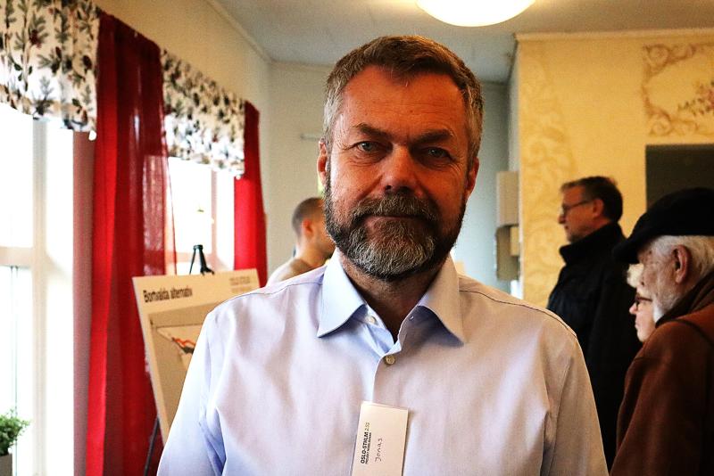 En man med skägg och blå skjorta står inomhus på vad som verkar vara ett evenemang eller en utställning, med andra människor och en anslagstavla i bakgrunden.