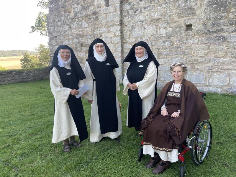 Fyra kvinnor i historiska eller religiösa kläder poserar framför en stenmur; en kvinna sitter i rullstol medan de andra står på gräset.