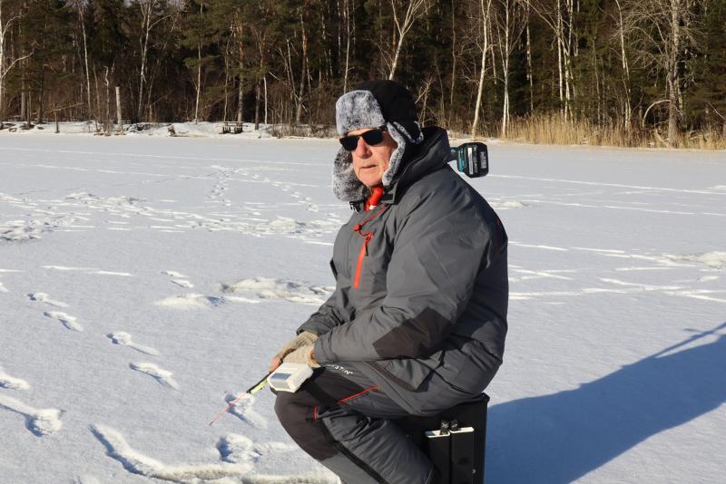 En äldre man i vinterkläder och solglasögon sitter på en låda och isfiskar på en frusen sjö med träd i bakgrunden.