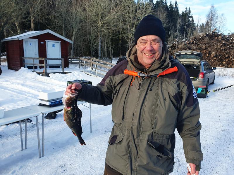 En vinterklädd man står utomhus på snö, ler och håller i en fisk. I bakgrunden syns bord, en bil och byggnader.