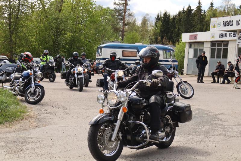 En grupp motorcyklister med hjälmar kör och parkerar sina motorcyklar nära en byggnad och en blå skåpbil på en delvis molnig dag.