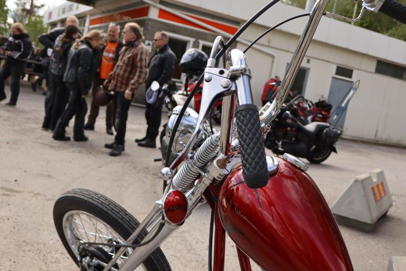 Närbild på en röd motorcykel parkerad utomhus, med en grupp människor som står och pratar i bakgrunden nära en byggnad.