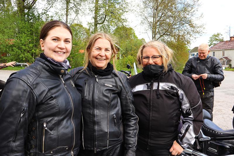 Tre kvinnor i svarta motorcykeljackor står tillsammans utomhus och ler mot kameran, medan en man i liknande utrustning står i bakgrunden och justerar något.