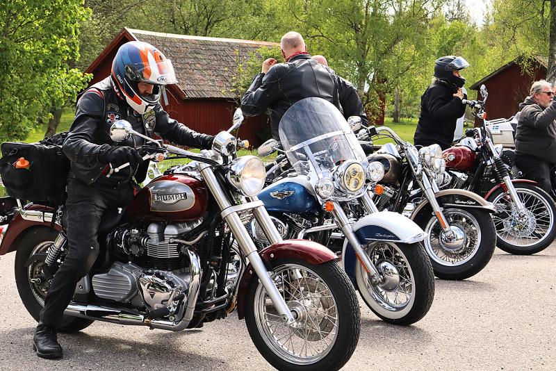 Flera motorcyklister i körutrustning står parkerade vid vägkanten med sina motorcyklar och förbereder sig för en tur; träd och röda träbyggnader syns i bakgrunden.