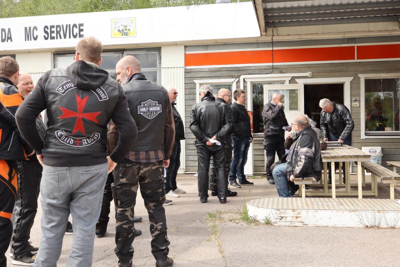 En grupp människor i läderjackor står och sitter utanför en byggnad märkt 